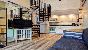 Ein kompakter Wohnraum mit Wendeltreppe, einer Küche mit weißen Schränken, einer Mikrowelle und einem Kühlschrank. Es gibt eine Couch mit gestreiften Kissen und einen Fernseher auf einem Schrank.