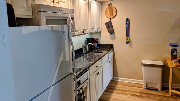 A compact kitchen with white cabinets, a refrigerator, a coffee maker, and a trash bin.