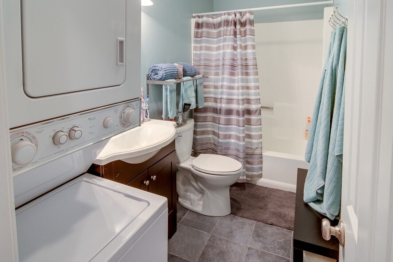Une petite salle de bain avec un lavabo, des toilettes et une baignoire, le tout de couleur blanche. On y trouve un rideau de douche à rayures et un porte-serviettes avec des serviettes.