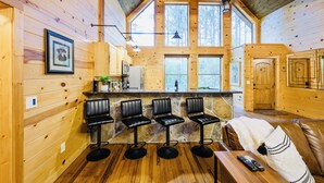 A cozy cabin interior with wooden walls, a stone accent wall, a bar area with stools, a sofa with a blanket, and large windows offering a view of the forest. - Candy Apple Cabin- Stay 3 get 4th Night FREE (Broken Bow)