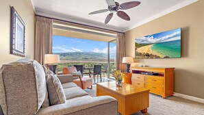 A hotel room with a sofa, a wooden coffee table, a TV, and a balcony with a view of the ocean and mountains.