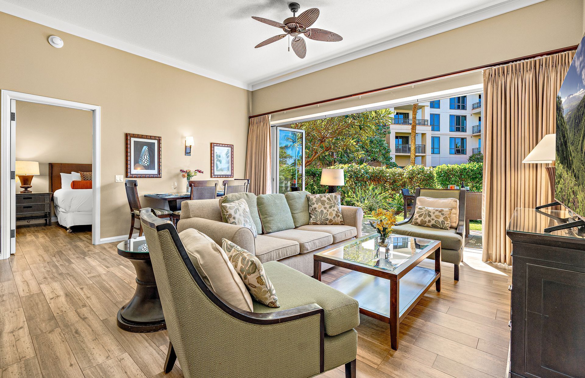 A spacious living room with a sofa, armchairs, and a coffee table. There is a dining area with a table and chairs, and a view of the outside through large windows.