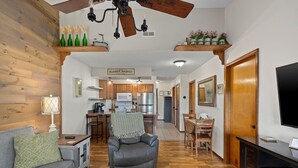 A cozy living room with a grey sofa, a leather armchair, and a TV stand. A kitchen with wooden cabinets and a dining area with a table and chairs are visible in the background. - Notch★The Chic Retreat★Trailer Parking1Mi to SDC (Branson West)