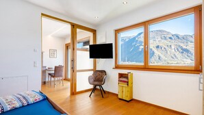 A bedroom with a bed, a desk, a chair, a TV, and a window with a mountain view. - Modern Apartment "3 Weger" with Panoramic Mountain Views, Balcony, and Wi-Fi (Andrian)