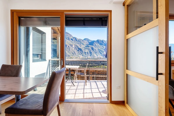 Una habitación con una mesa y sillas de madera, una puerta corredera de vidrio que da a un balcón con vista a la montaña.