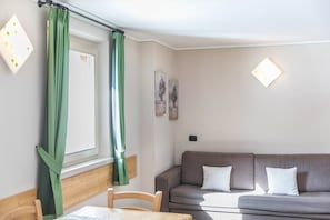 Une pièce avec une tête de lit en bois, un canapé marron avec des coussins blancs, une table en bois et une fenêtre ornée de rideaux verts.