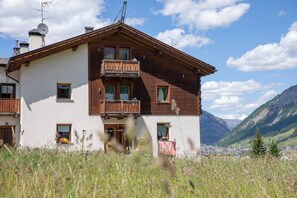 A two-story wooden house with a balcony, a satellite dish, and a mountain backdrop.