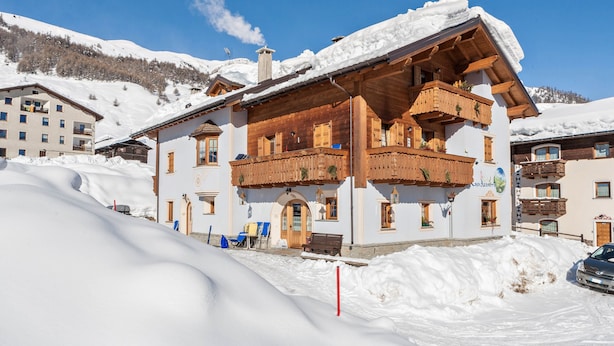 A snow-covered building with wooden balconies, a clear sky, and a snowy mountain in the background. - Casa Azzurra Piano Terra with Mountain View, Wi-Fi & Garden; Parking Available (Livigno (SO))