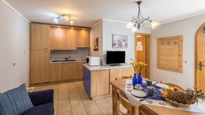 A compact kitchen with wooden cabinets, a dining table with a fruit basket, and a TV on the counter.