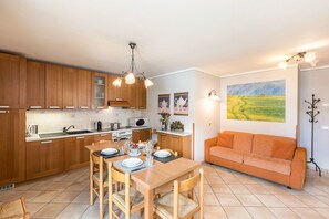 A modern kitchen with wooden cabinets, a dining table with chairs, and a sofa in a living area with a painting on the wall.