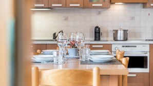 Mesa lista para comer con platos, vasos y un pequeño centro de mesa con planta en una cocina con alacenas de madera y estufa.