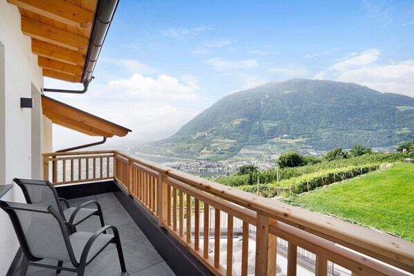 Un balcón con barandilla de madera, dos sillas y vistas a un paisaje montañoso.