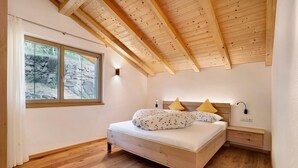 Een slaapkamer met een houten plafond, een bed met kussens, een nachtkastje en een raam met uitzicht op rotsen.
