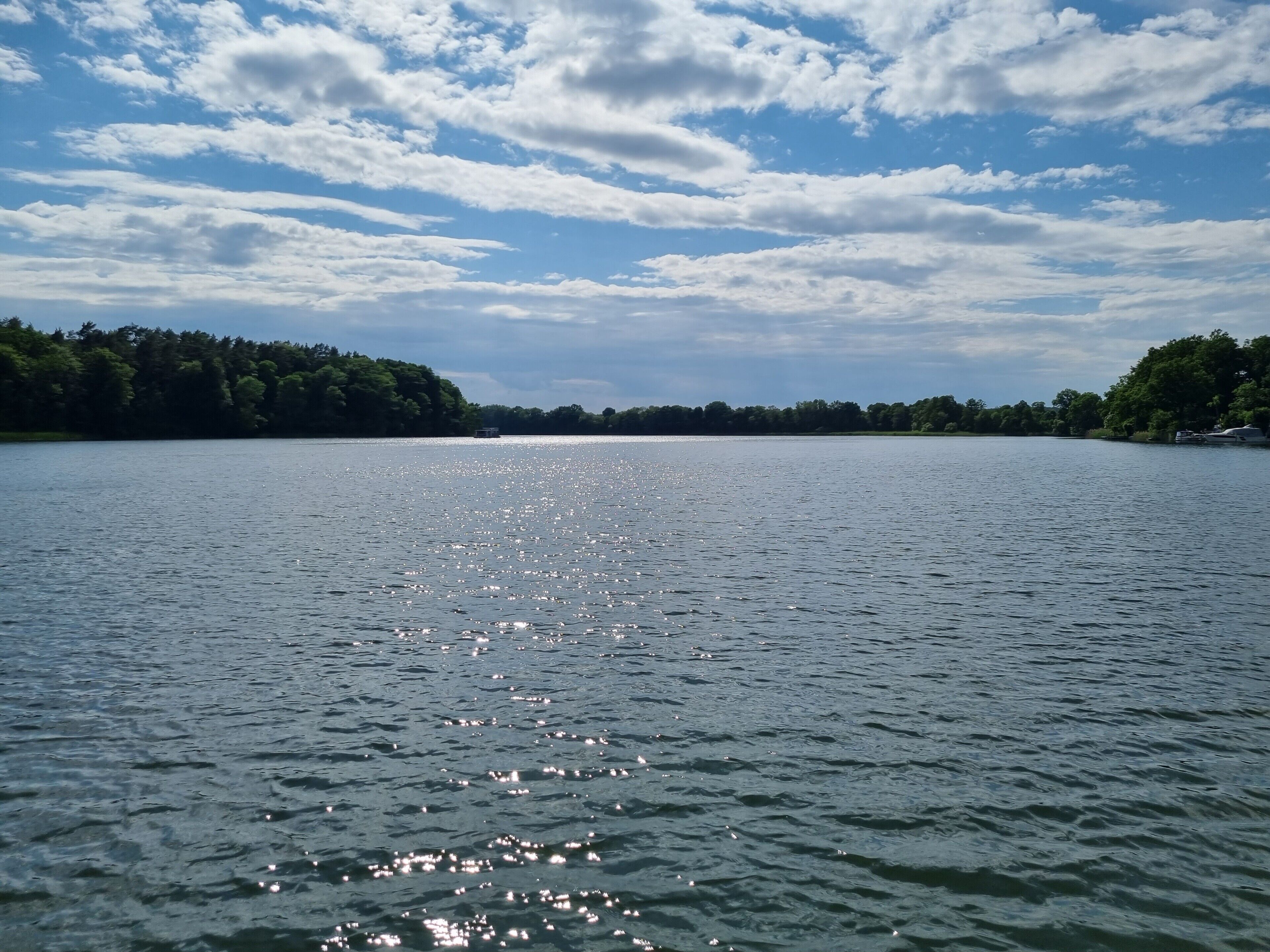 Un lac paisible, avec un bateau visible au loin, entouré d’une forêt dense et sous un ciel partiellement nuageux.