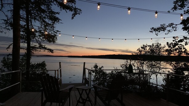 Terraza de madera con sillas y luces colgantes con vista a una masa de agua tranquila al atardecer.
