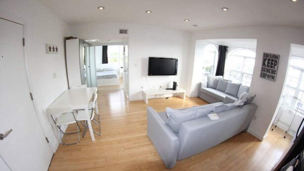 Un salon moderne avec un canapé gris, une table à manger blanche avec des chaises, un téléviseur à écran plat fixé au mur et une grande fenêtre laissant entrer la lumière naturelle.