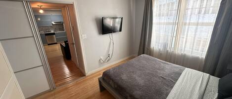 Une chambre à coucher comprenant un lit, un téléviseur fixé au mur, une fenêtre avec des rideaux et une porte donnant sur une salle de bain.
