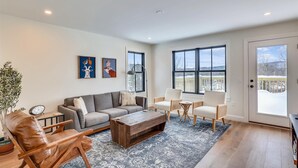 A spacious living room with a grey sofa, wooden coffee table, and a blue patterned rug. There are two framed artworks on the wall, a potted plant, and a large window with a view of a snowy deck. - Village with a View: 4 br, a/c, deck, Mansfield views! (Stowe)