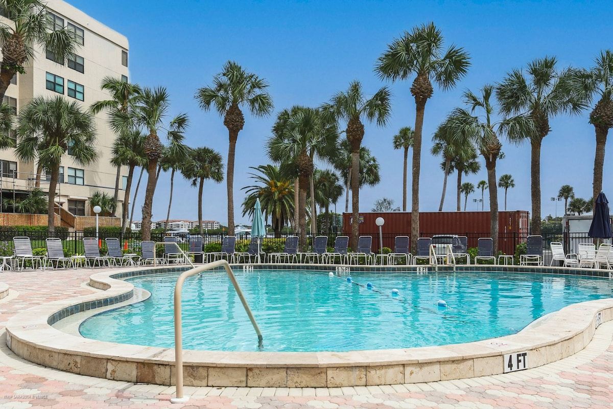 A swimming pool with a curved edge, surrounded by palm trees and lounge chairs.