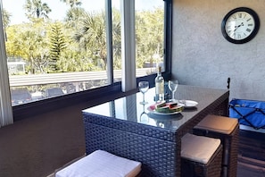 A dining area with a glass-top table, wicker chairs, a clock on the wall, and a view of palm trees outside. - Renovated 3BR Harbor Towers: Pools, Tennis & Beach (Siesta Key)