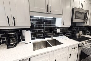 A kitchen with white cabinets, a black tiled backsplash, a stainless steel sink, a coffee maker, and a microwave. - Renovated 3BR Harbor Towers: Pools, Tennis & Beach (Siesta Key)