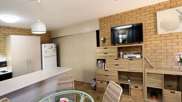 Een moderne keuken met een flatscreen televisie aan een bakstenen muur, houten kasten en een eethoek met rieten stoelen.