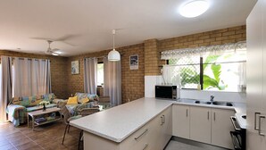 Een kleine keuken met een centraal kookeiland, een eethoek met een bank en stoelen, en uitzicht op groen via een raam.