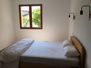 A single bed with a wooden headboard, a white mattress, and a white blanket. There is a window with a wooden frame and a view of greenery outside.