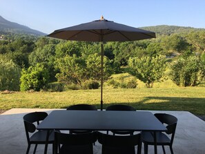 A patio table with four chairs and a large umbrella, set against a backdrop of a lush green landscape and distant hills.