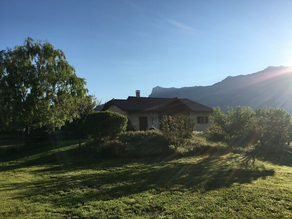 A house with a sloped roof, surrounded by trees and grass, with mountains in the background.