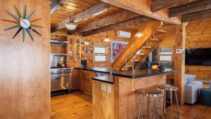 Interior rústico de una cabaña con zona de cocina, vigas de madera, un taburete de bar y un reloj en la pared.
