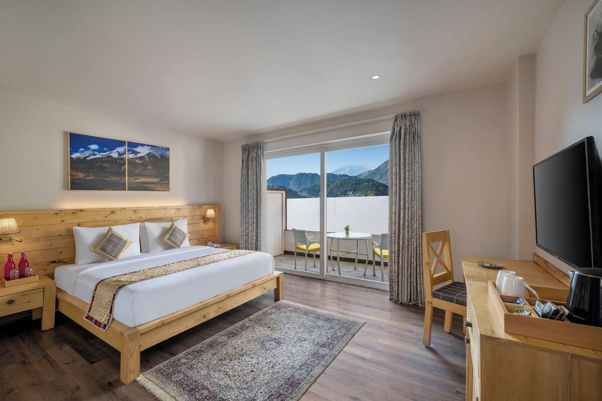 Ein Hotelzimmer mit einem hölzernen Bett, einem Fernseher, einem Schreibtisch mit Stuhl und einem Balkon mit Bergblick.