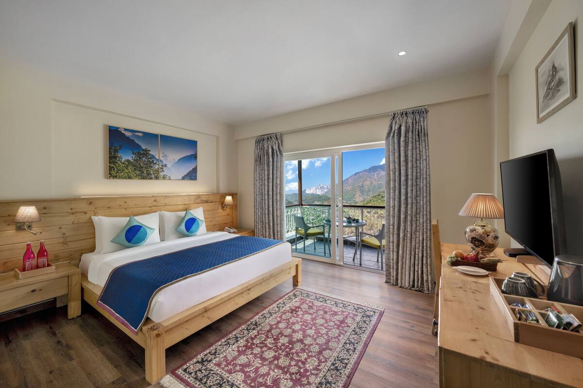 Ein Hotelzimmer mit einem hölzernen Bett, einem großen Fenster mit Vorhängen, einem Fernseher und einem Balkon mit Tisch und Stühlen.