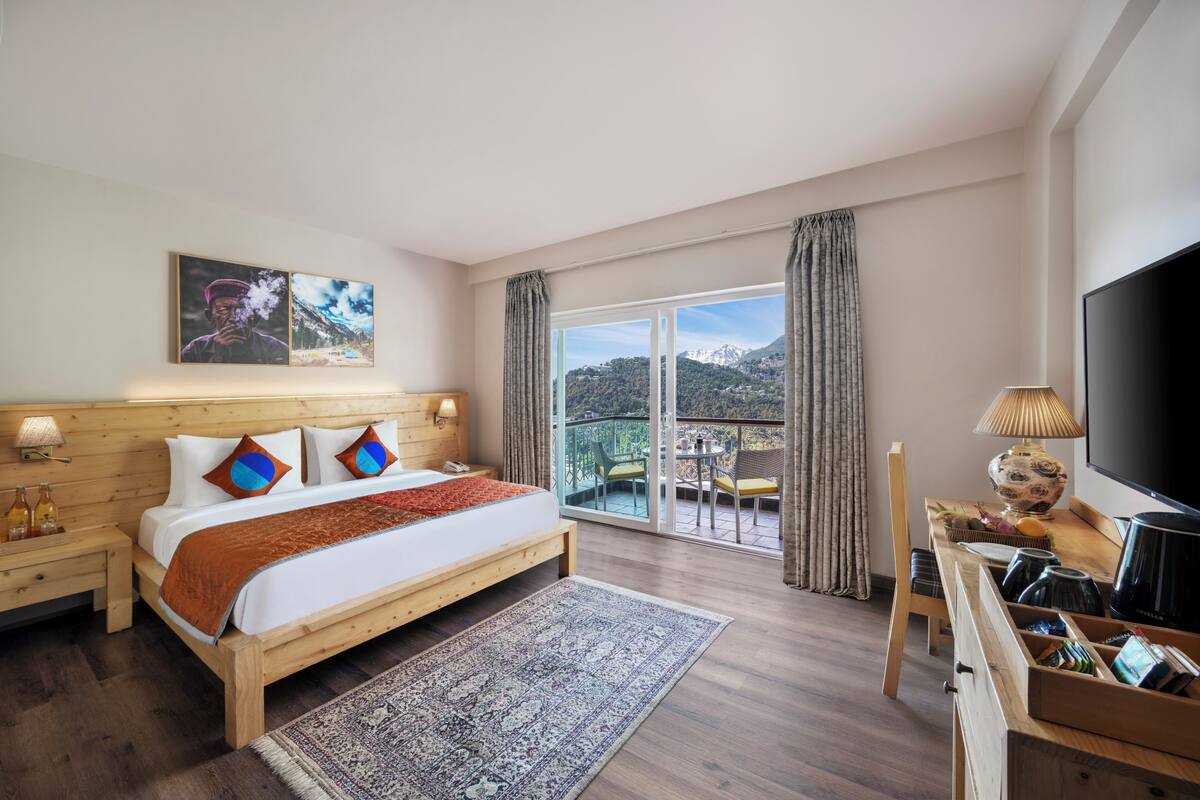 Ein Hotelzimmer mit einem hölzernen Bett, einem Fernseher, einem Balkon mit Bergblick und einer Essecke mit Tisch und Stühlen.
