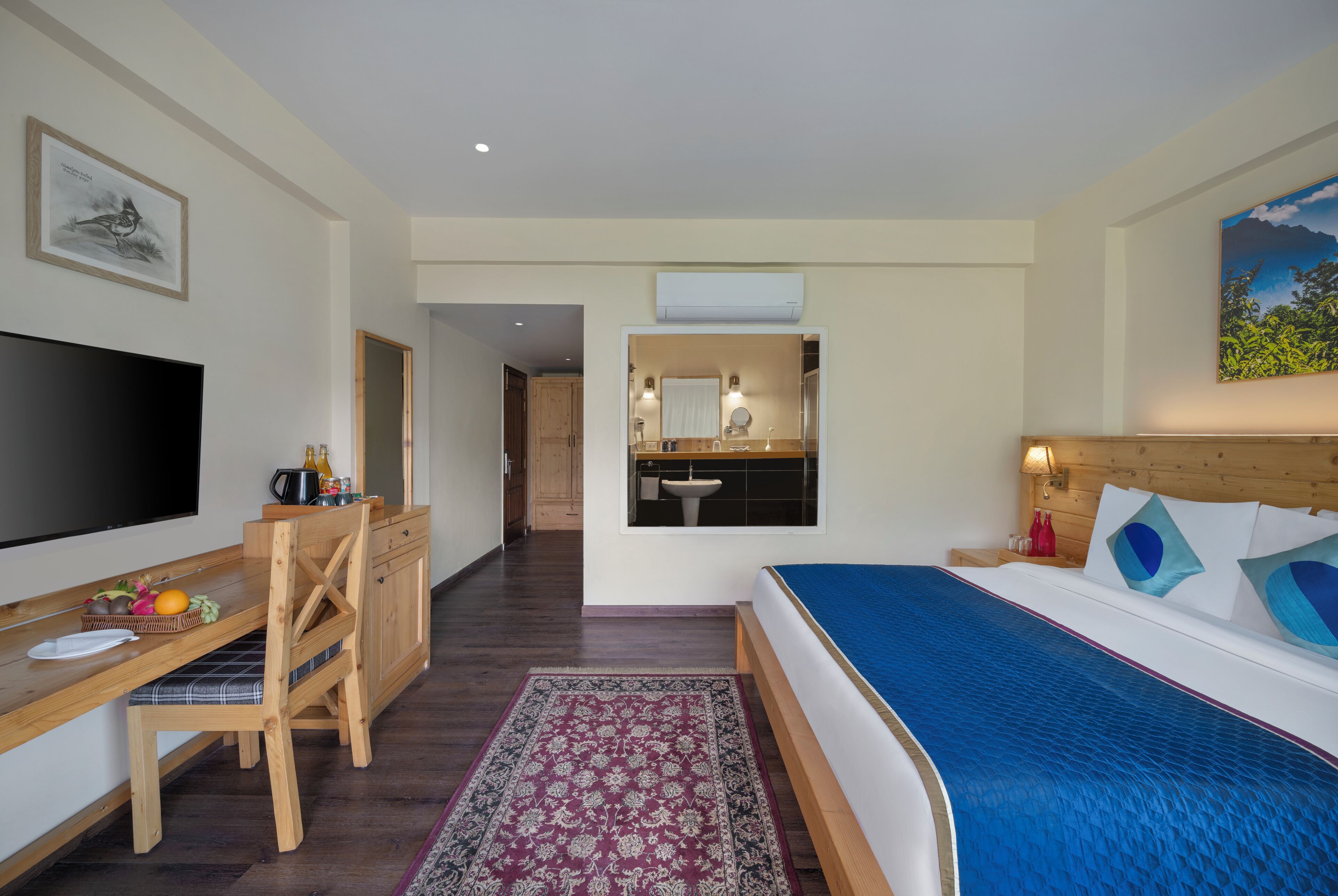 A hotel room with a bed, a desk with a flat-screen TV, a wooden chair, a small dining table with fruits, a patterned rug, a bathroom with a sink and mirror, and a painting on the wall.