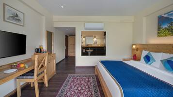 A hotel room with a bed, a desk with a flat-screen TV, a wooden chair, a small dining table with fruits, a patterned rug, a bathroom with a sink and mirror, and a painting on the wall.