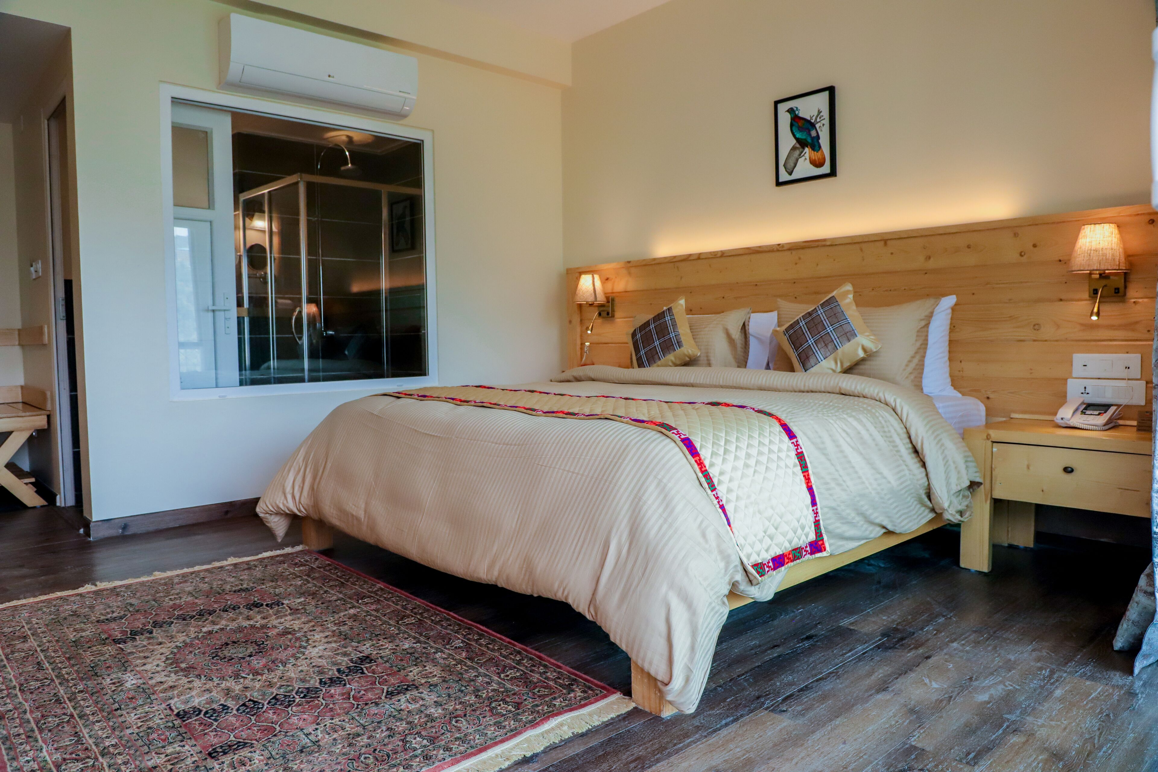 A bedroom with a large bed, a nightstand with a lamp, a wooden headboard, a window with a view, and a wall-mounted air conditioner.