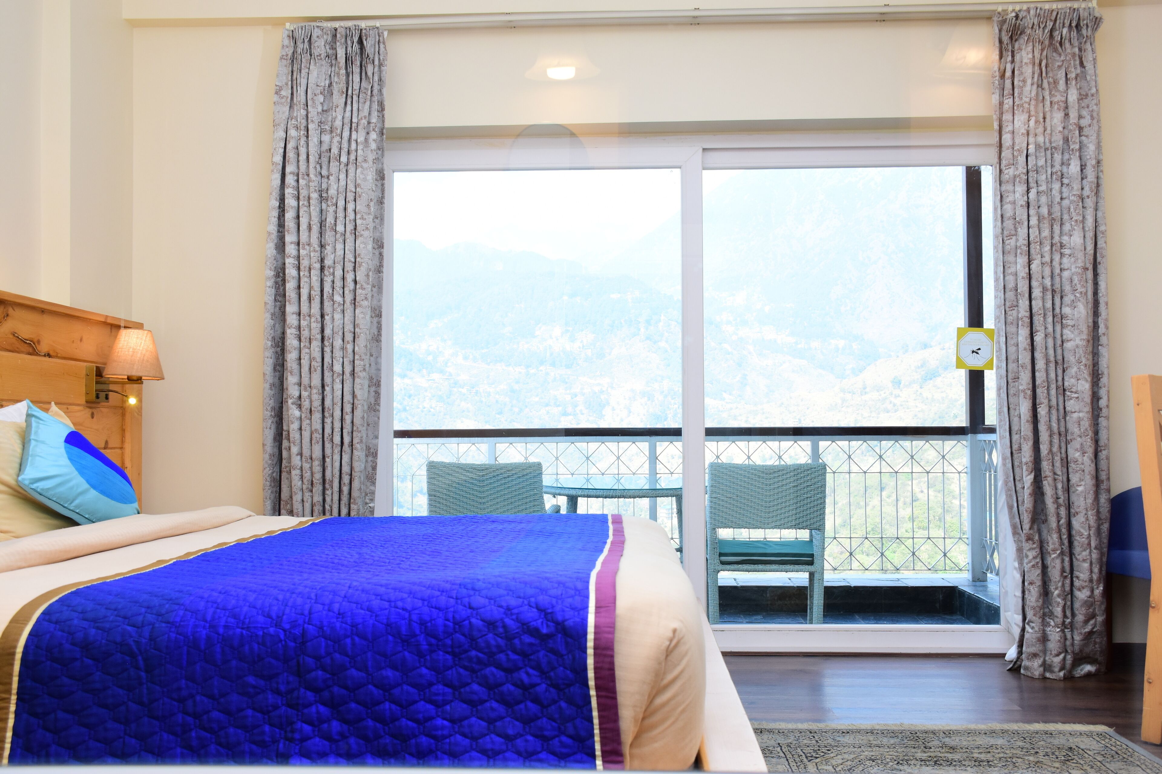 Un dormitorio con cama, mesita de noche, lámpara, ventana con cortinas y vista al exterior.
