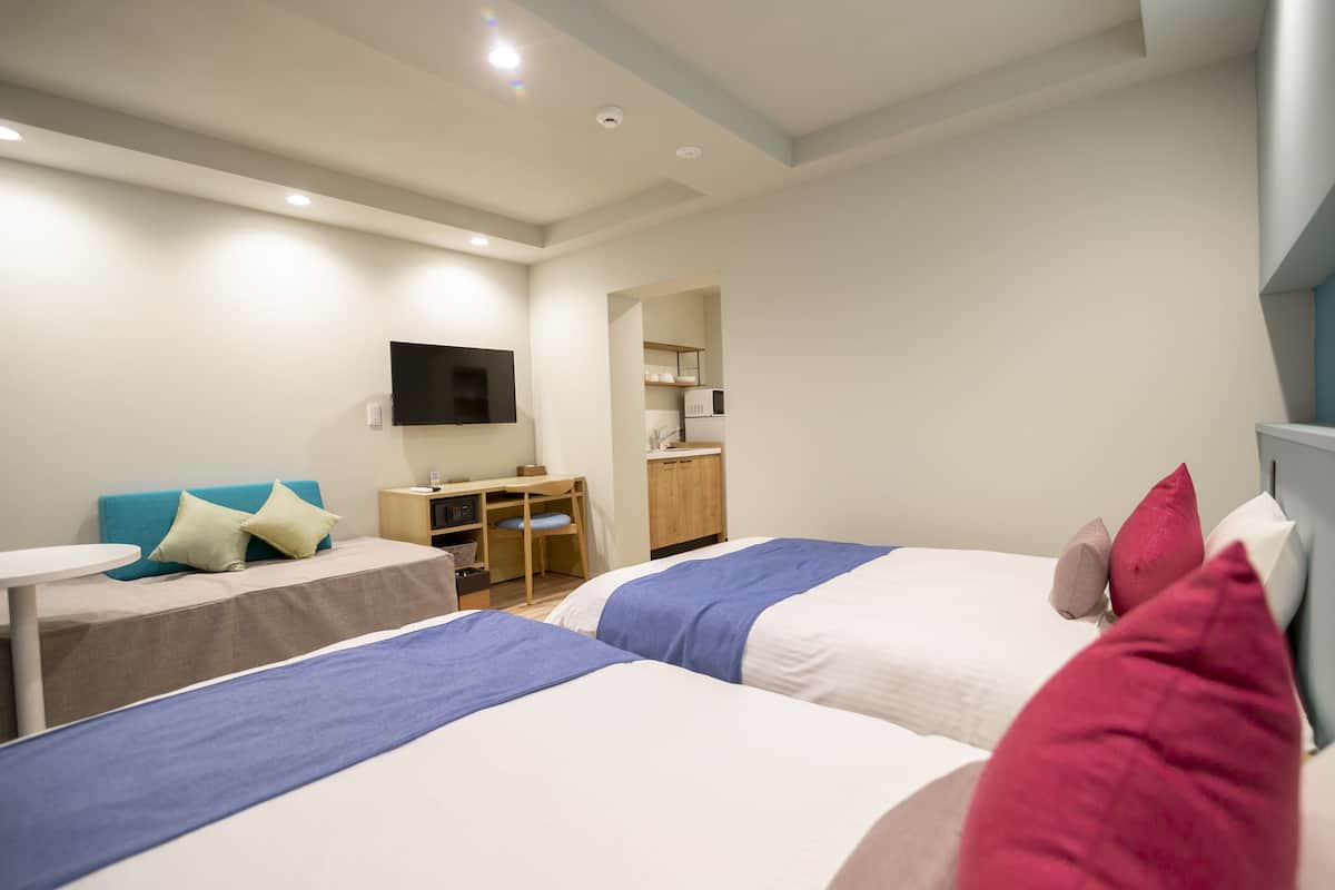 A hotel room with two beds, a desk, a chair, a TV, and a kitchenette.