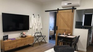 A modern bedroom with a bed, a TV, a wooden dresser, a desk with chairs, and a sliding door leading to a bathroom.