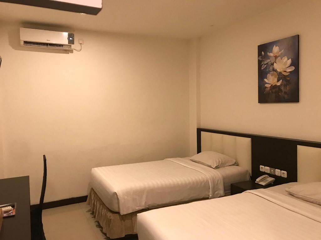 Une chambre d’hôtel avec deux lits simples, un bureau, une chaise, un appareil de climatisation, un tableau et un téléphone sur la table de chevet.