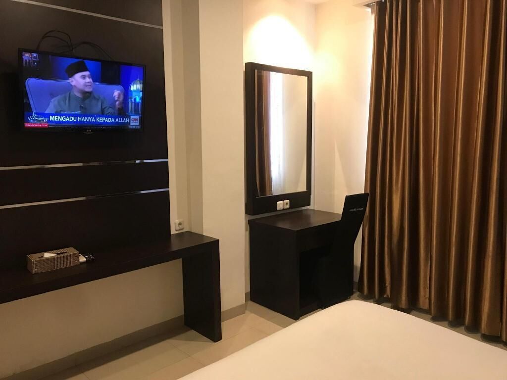 Hotellihuone, jossa on televisio, jolta näkyy uutiskanava, suuri peili, työpöytä ja sänky.