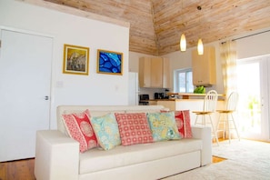 Un salón acogedor con un sofá blanco decorado con cojines estampados, techo de madera y una zona de cocina al fondo.