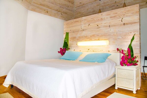 Una camera da letto con soffitto in legno, un letto con biancheria bianca, due cuscini blu, un comodino con una lampada e un bouquet di fiori.