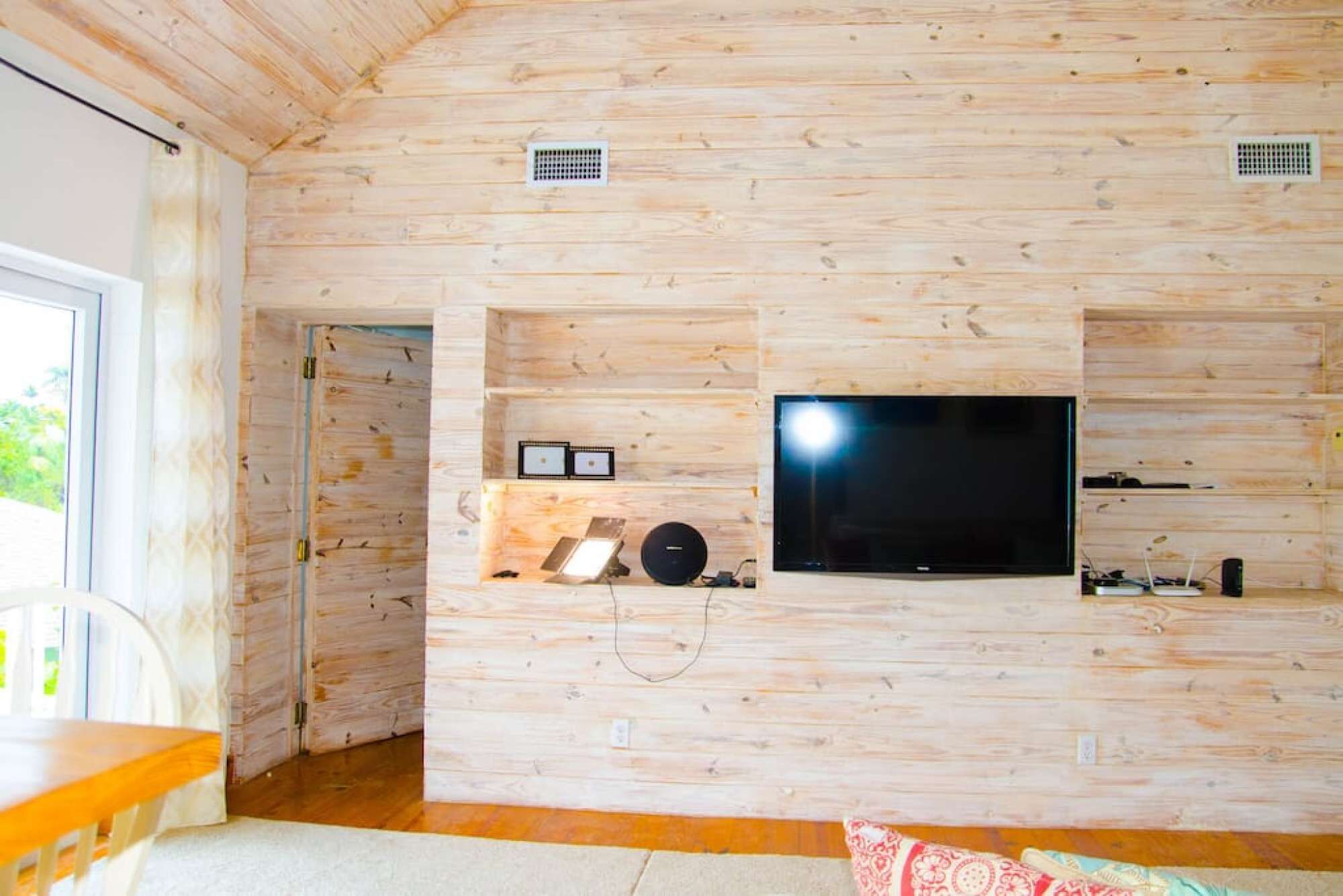 Een kamer met houten lambrisering, een flatscreen televisie aan de muur, een klein plankje met decoratieve objecten en een raam dat voor daglicht zorgt.