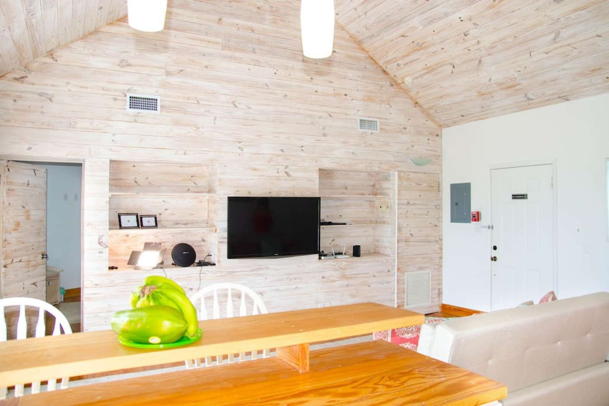 A wooden interior with a dining table, chairs, a sofa, a TV mounted on the wall, and a door with a sign.