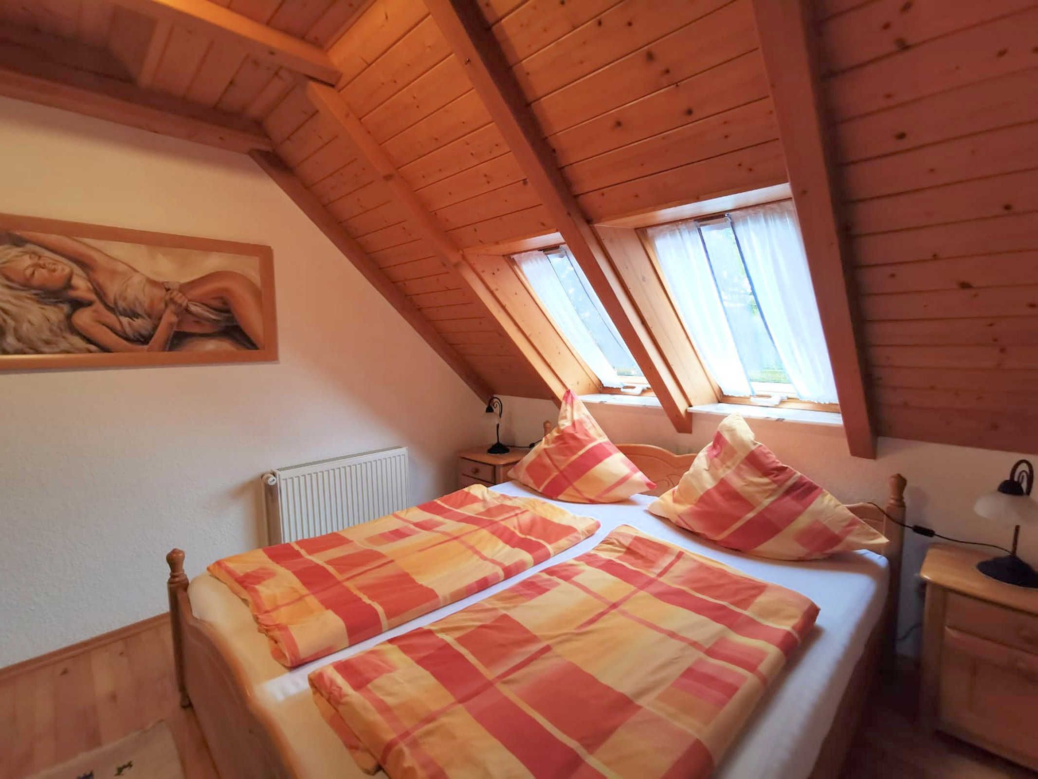 Kodikas ullakkohuone, jossa on kalteva katto, puiset kattopalkit, suuri sänky ruutukuosisella vuodevaatteella, patteri ja seinällä oleva taulu.