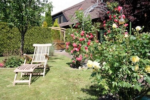 Ein Garten mit einem Holzsessel, einem Rosenbusch und einem Haus mit einem Ziegeldach.