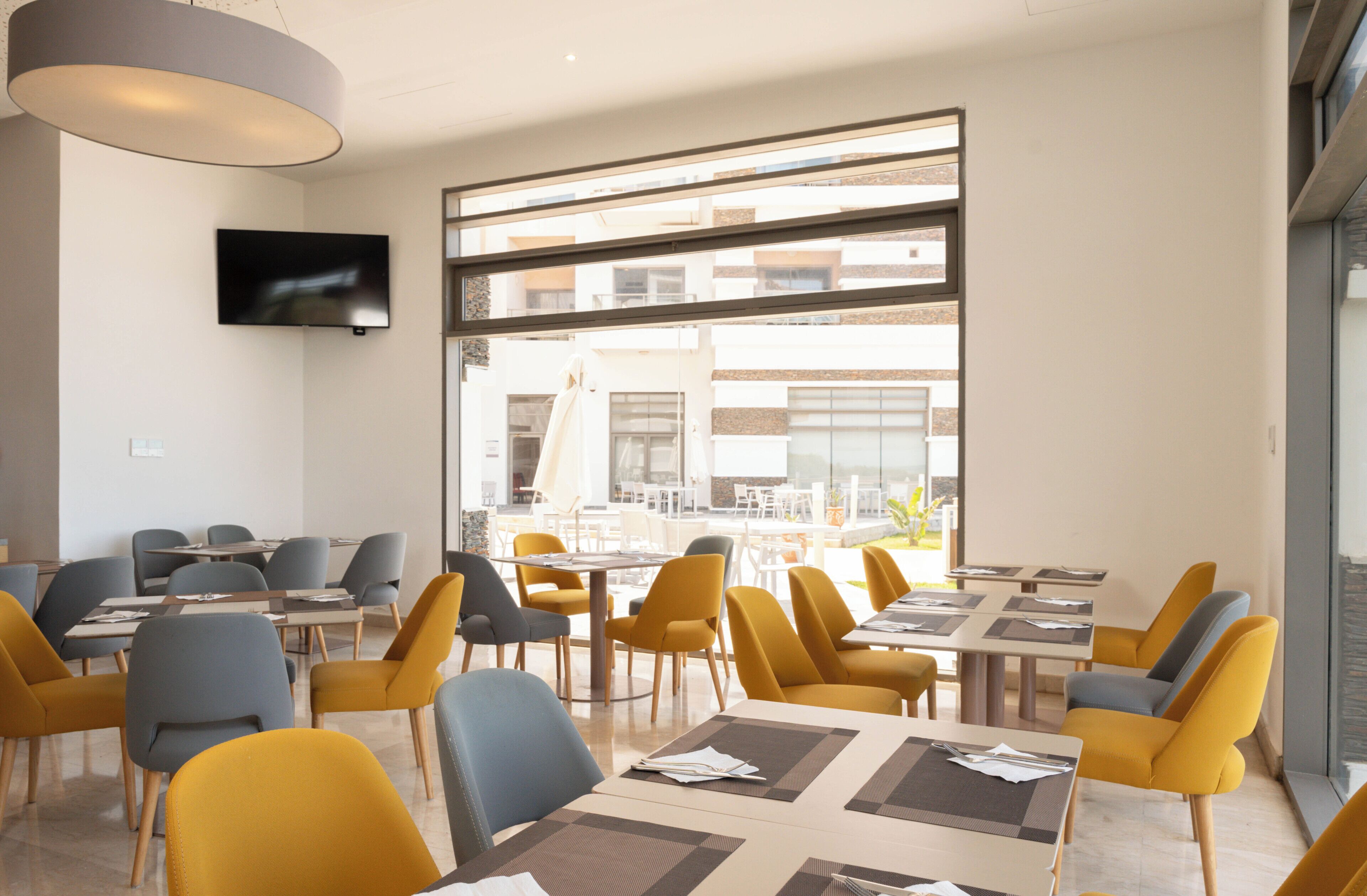 Un comedor moderno con mesas y sillas, un televisor de pantalla plana en la pared y ventanas amplias con vistas al exterior.