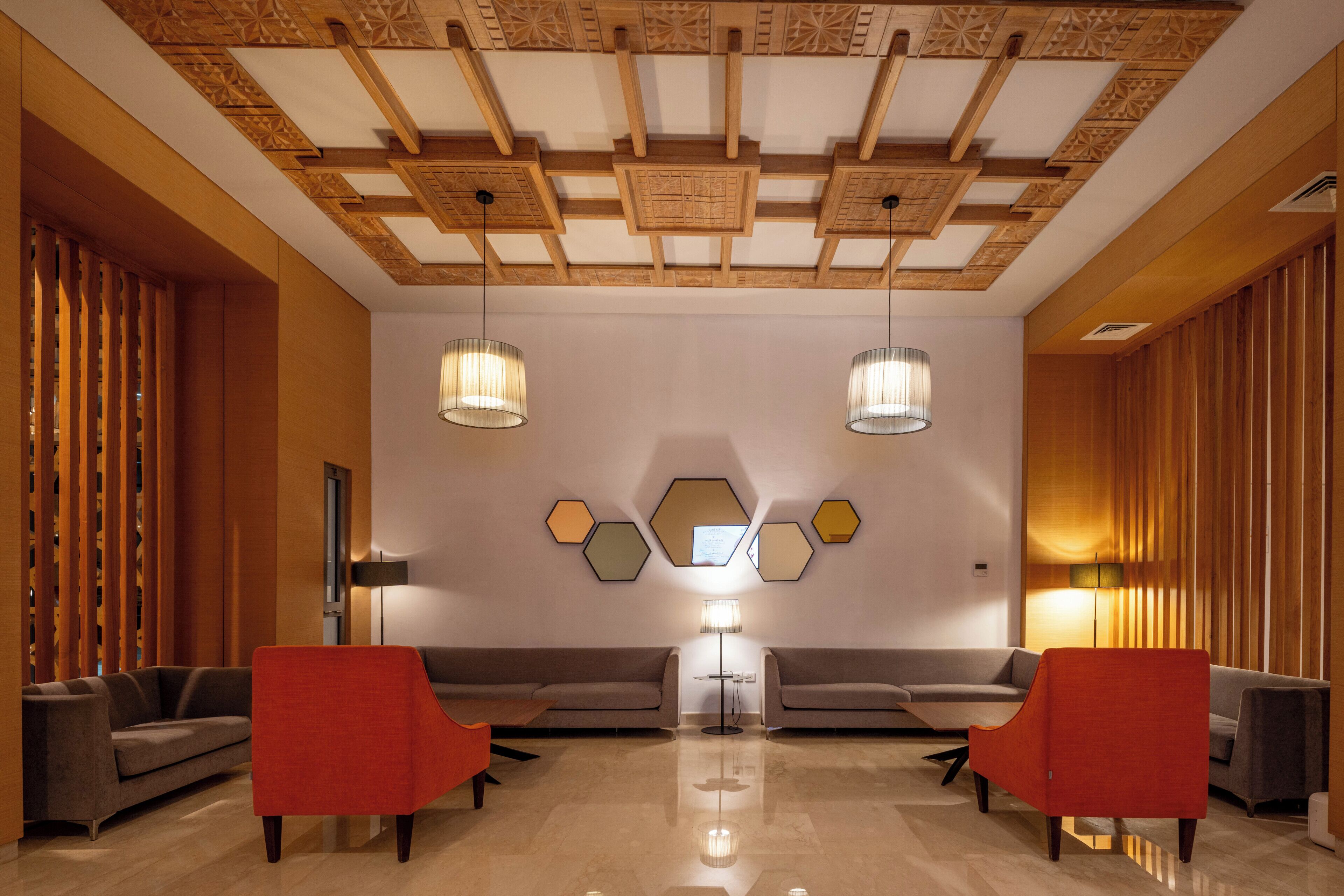 A modern lobby with a geometric ceiling, two sofas, a red armchair, and a floor lamp.